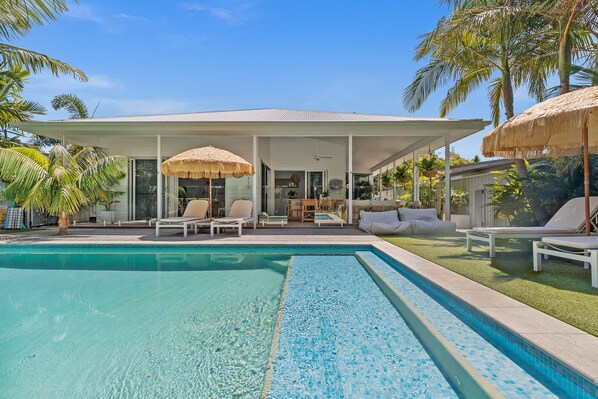 Un espace aménagé au bord de la piscine, comprenant un coin salon couvert, des chaises longues et un parasol en chaume.