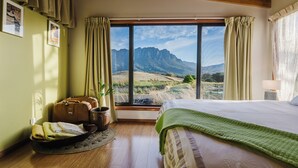A bedroom with a large window, wooden flooring, a bed with a green blanket, a potted plant, and a suitcase.