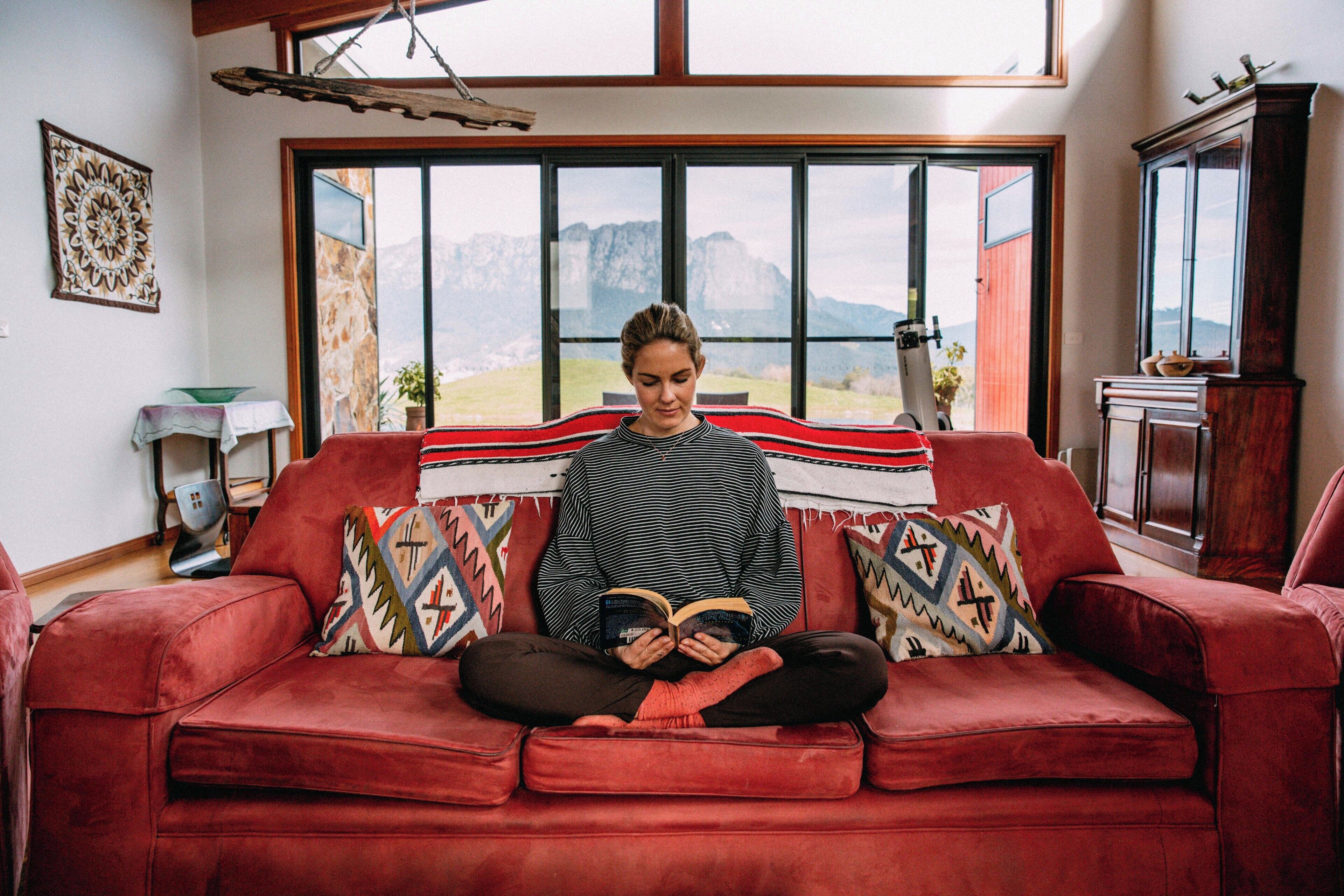 Une personne est assise sur un canapé rouge, un livre à la main, dans une pièce dotée d’une grande fenêtre, d’un meuble en bois et d’un élément décoratif mural.