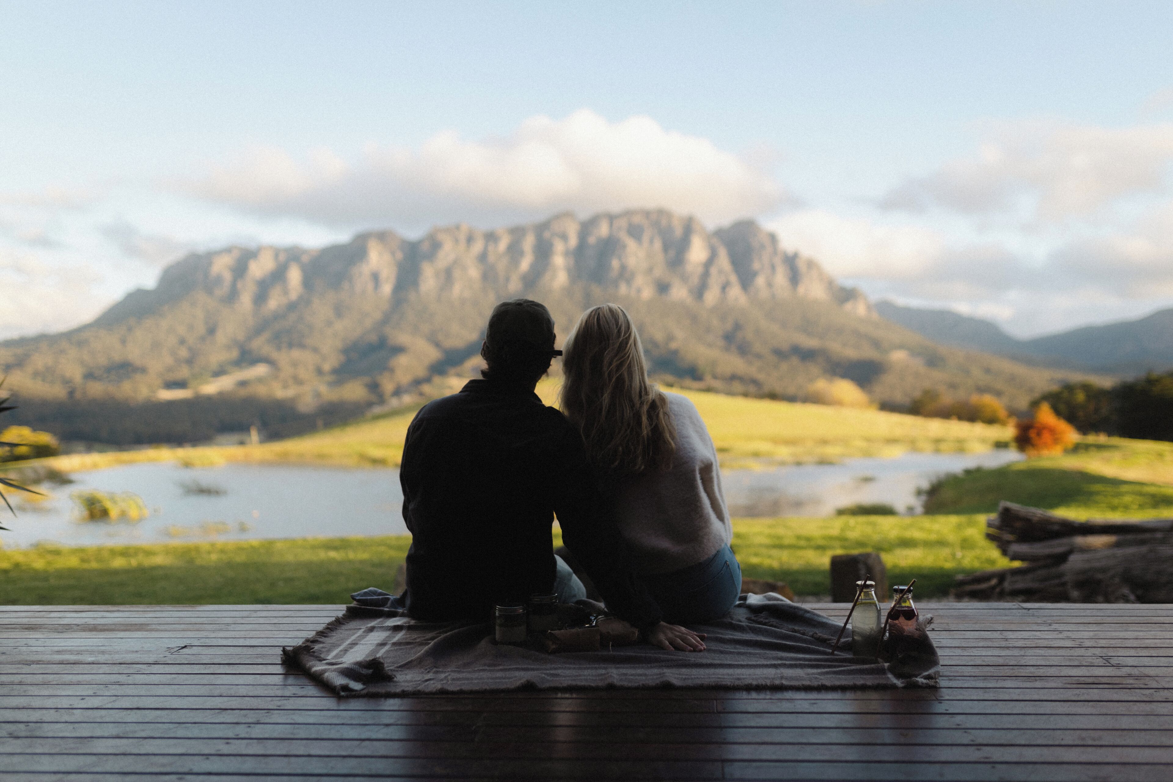 A couple sitting on a wooden deck overlooking a lake with mountains in the background.