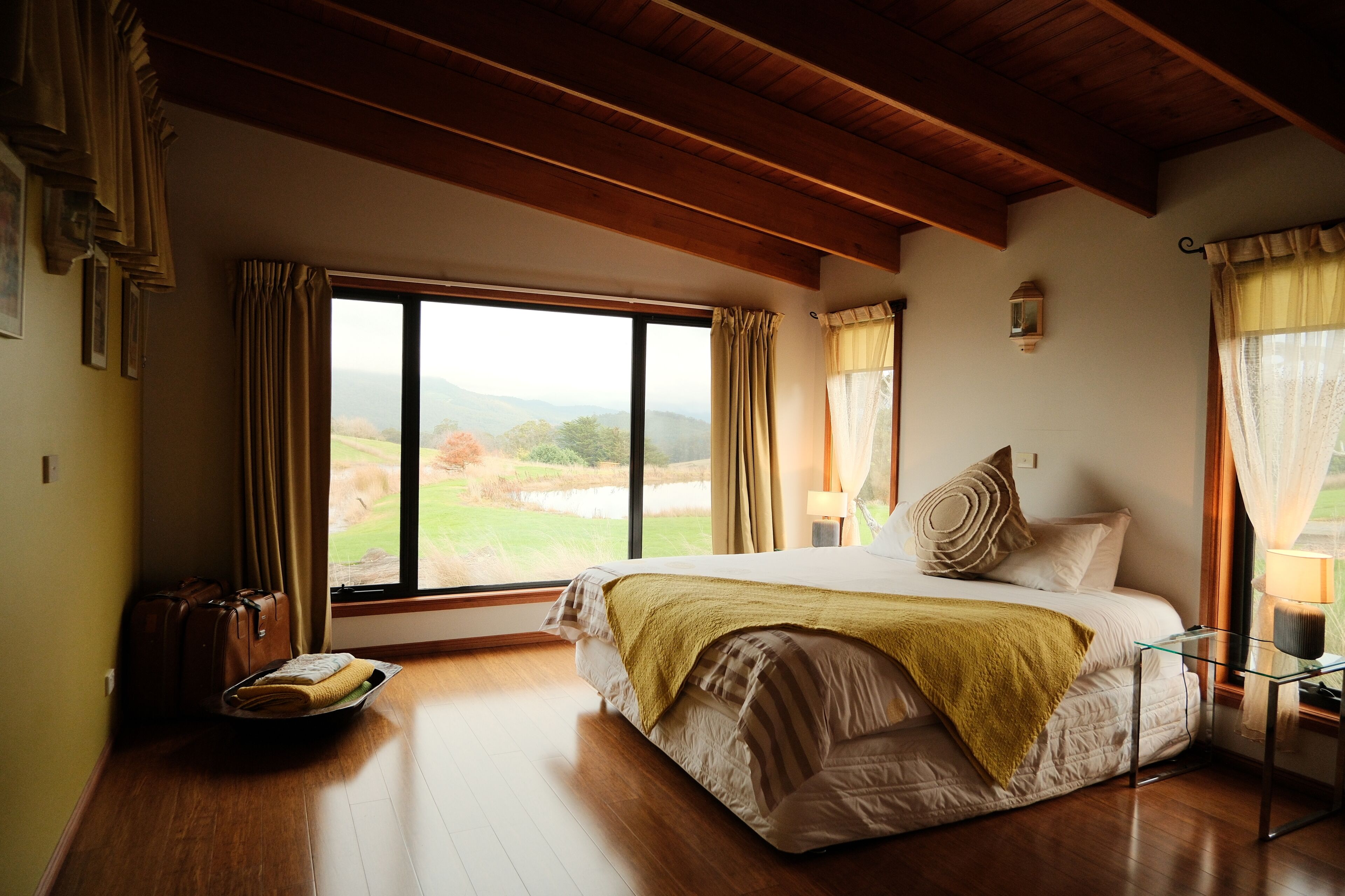 Une chambre avec une grande fenêtre, des poutres en bois, un lit recouvert d’un plaid jaune et une valise posée sur le sol.