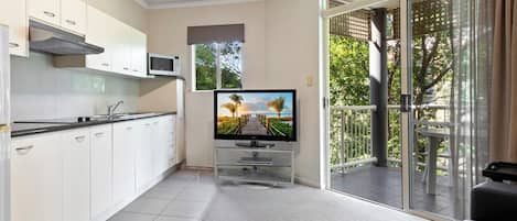 Una cocina moderna con armarios blancos, un microondas y un televisor sobre un mueble bajo. Hay una puerta corrediza de vidrio que da a un balcón con vistas a la vegetación.