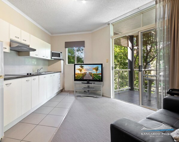 A modern kitchen with white cabinets, a microwave, and a TV on a stand. There is a sliding glass door leading to a balcony with a view of greenery.