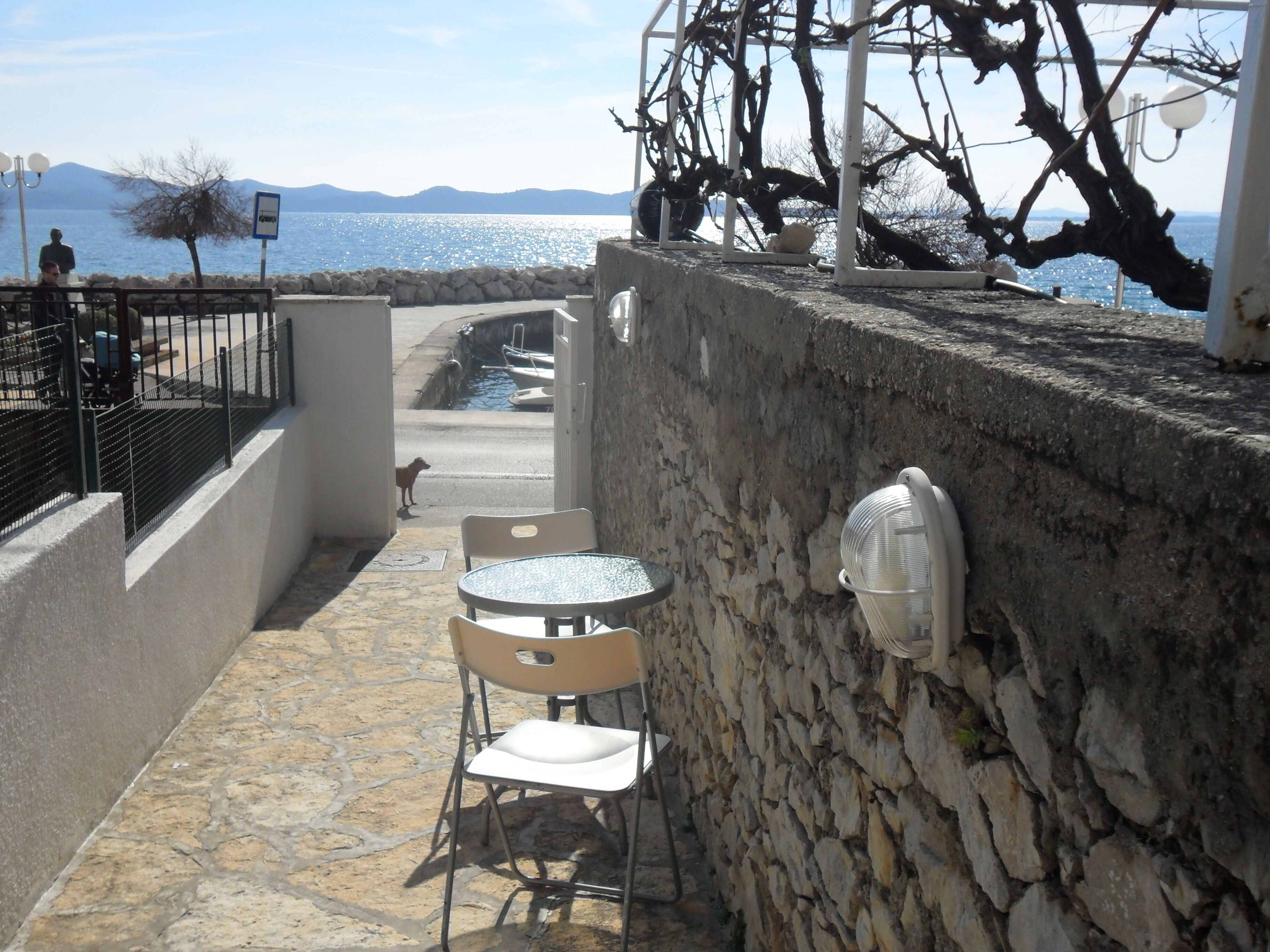 A stone-walled outdoor seating area with a table and chairs, overlooking a body of water and mountains.