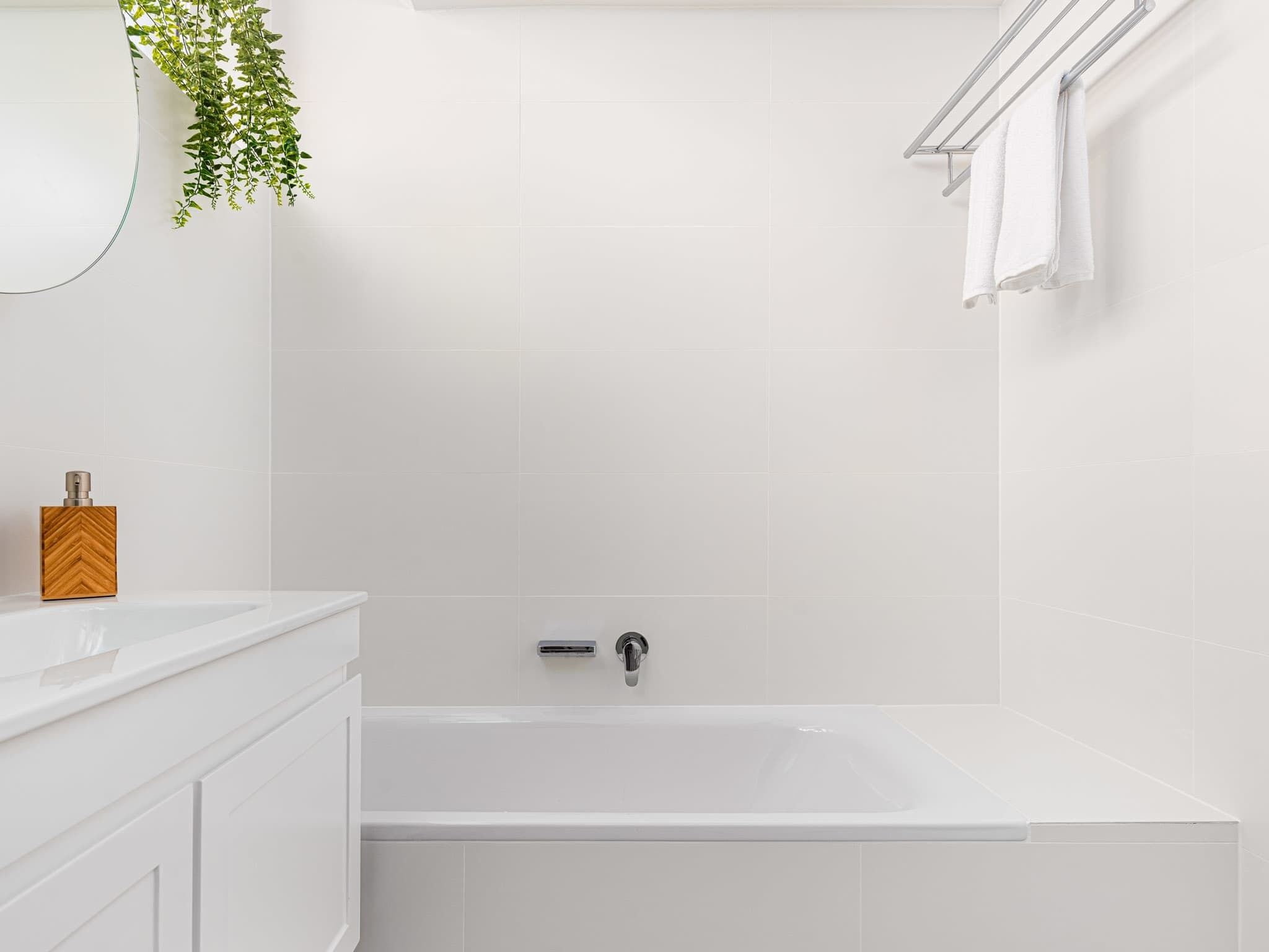 Un baño moderno con azulejos blancos, una bañera independiente, un dosificador de jabón de madera y una planta colgante.