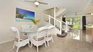 Un comedor con una mesa de vidrio y sillas de mimbre blanco, un ventilador de techo, una escalera con barandas blancas y un cuadro de una escena de playa.