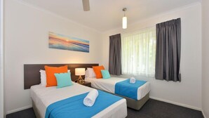 Habitación de hotel con dos camas, cada una con colcha azul y ropa de cama blanca, una mesita de noche con lámpara, un cuadro en la pared y una ventana con cortinas.