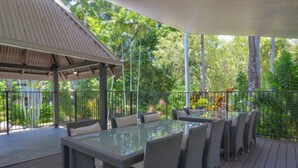 Un espacio comedor al aire libre con techo, mobiliario de mimbre y una mesa con superficie de vidrio, rodeado de abundante vegetación.