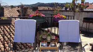 A rooftop terrace with two lounge chairs, a small table with a bottle and glasses, and potted plants.