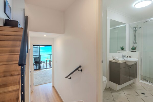 Et moderne badeværelse med brusebad, toilet og håndvask. Der er monteret et gelænder på væggen, og en glasdør fører ud til en balkon med udsigt.