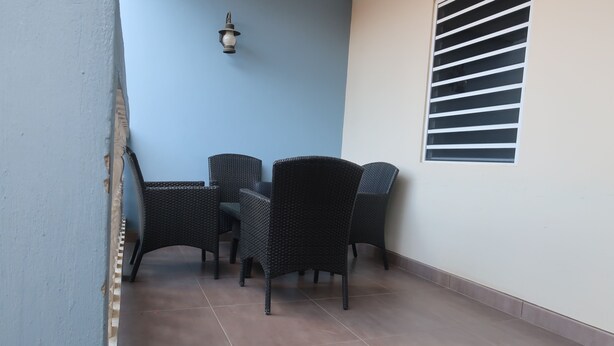 A corner of a room with a blue wall, a window with horizontal blinds, and a table with four wicker chairs. - Two Bedroom two bathroom Apartment A ground level (Willemstad)