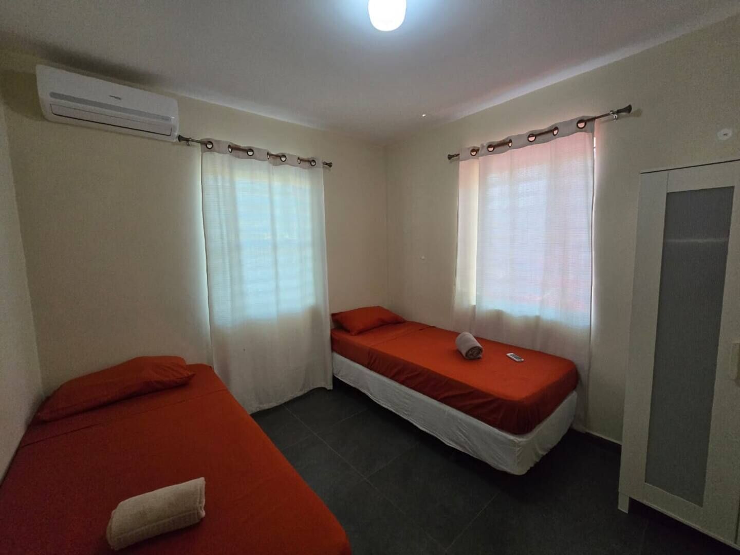 A small, simple room with two single beds, a wardrobe, and an air conditioning unit.
