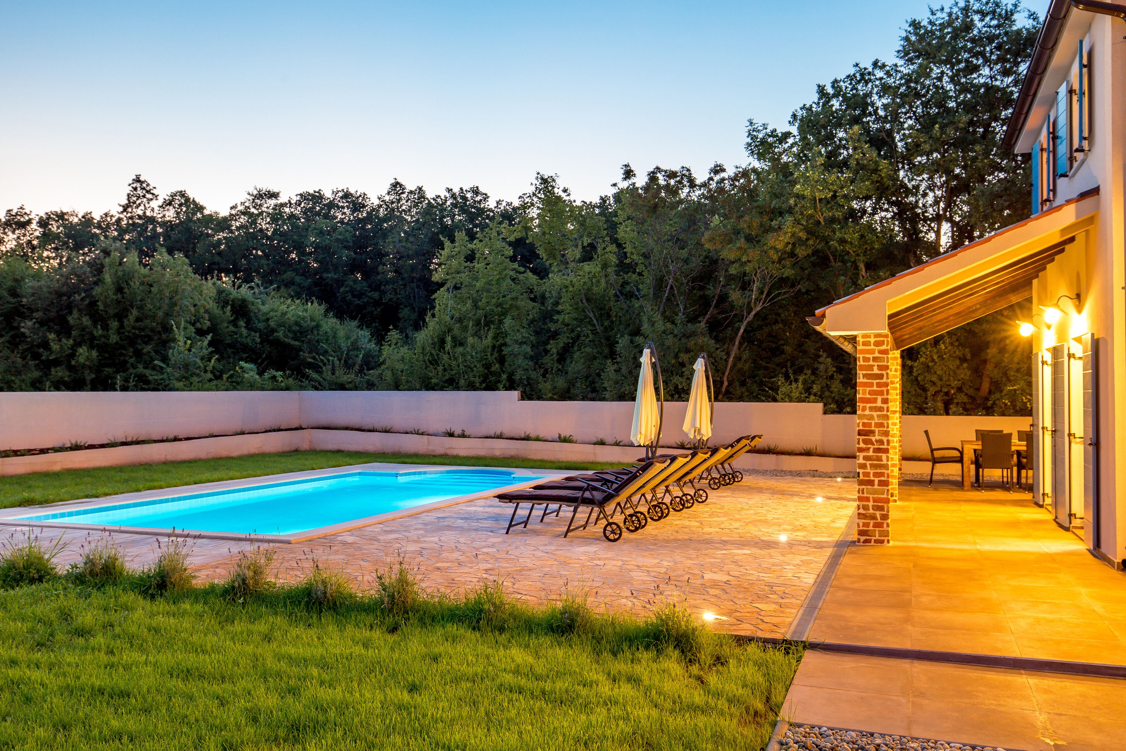 A swimming pool with lounge chairs and umbrellas, a patio area with a table and chairs, and a brick wall surrounding the space.