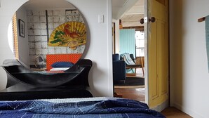 Ein Schlafzimmer mit einem Bett, einem runden Spiegel, einer Wand mit einer Dekoration und Blick in einen weiteren Raum.