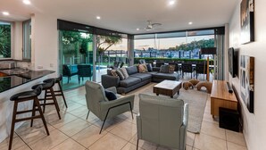 Una sala moderna con un sofá grande, una mesa de centro y un comedor con sillas. Hay una zona de cocina con una barra y taburetes. La habitación tiene ventanas amplias que ofrecen vistas a zonas verdes y al puerto deportivo.