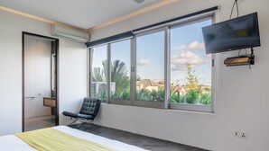 Une chambre d’hôtel moderne, dotée d’un grand lit, d’un fauteuil, d’une télévision et d’une fenêtre donnant sur un paysage verdoyant et des bâtiments.