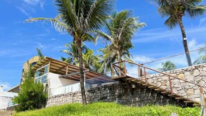 Una casa moderna con una escalera de piedra que conduce a una terraza de madera, rodeada de abundante vegetación y palmeras.