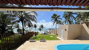 Zona de piscina con vista al océano, palmeras y un porche cubierto.