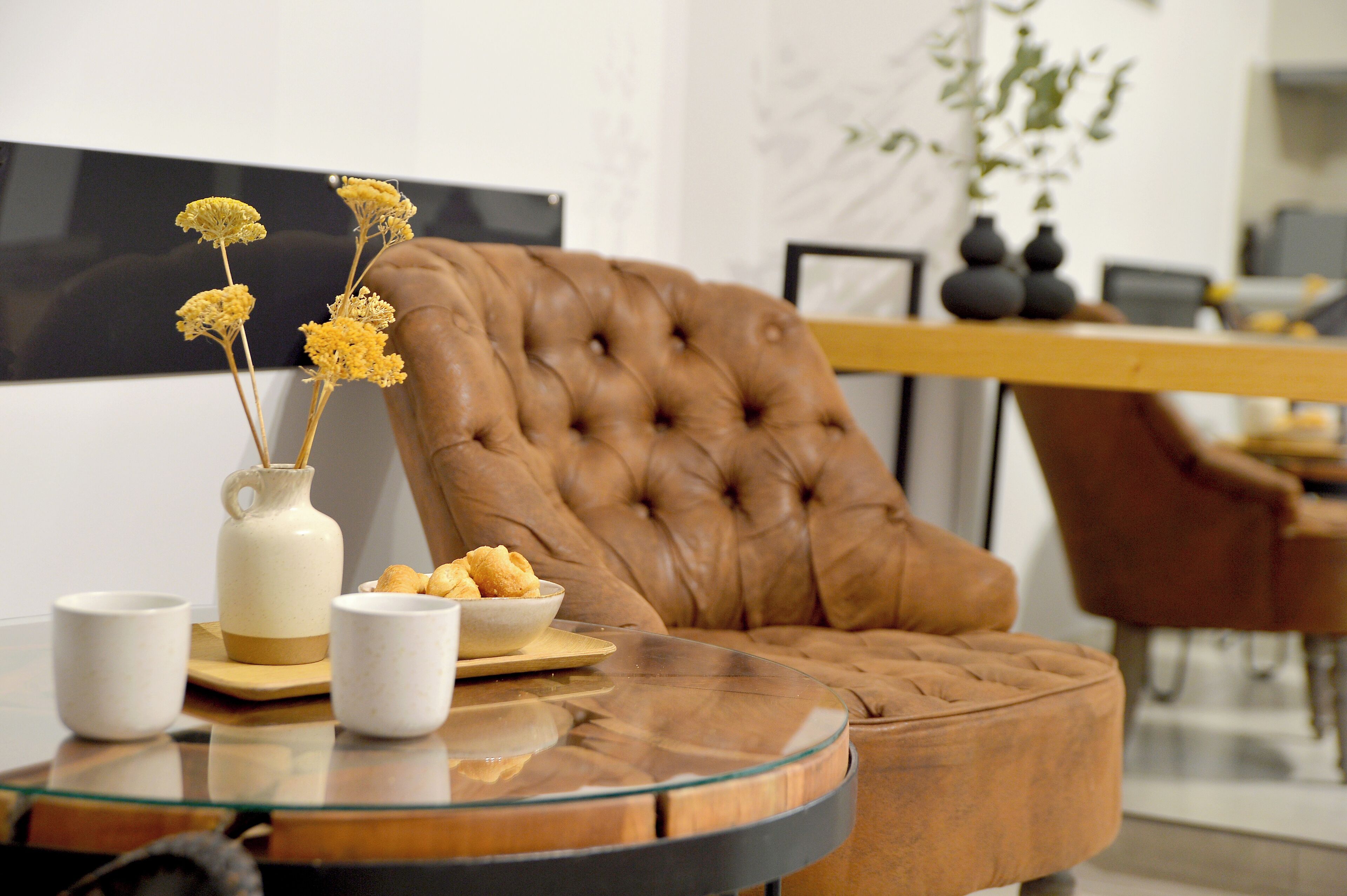 A modern living room with a tufted leather armchair, a glass coffee table, and a vase with dried flowers.