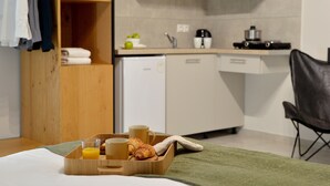 Una camera d'albergo con un letto in legno, un vassoio della colazione con croissant e succo di frutta, una coperta verde e una piccola cucina sullo sfondo.