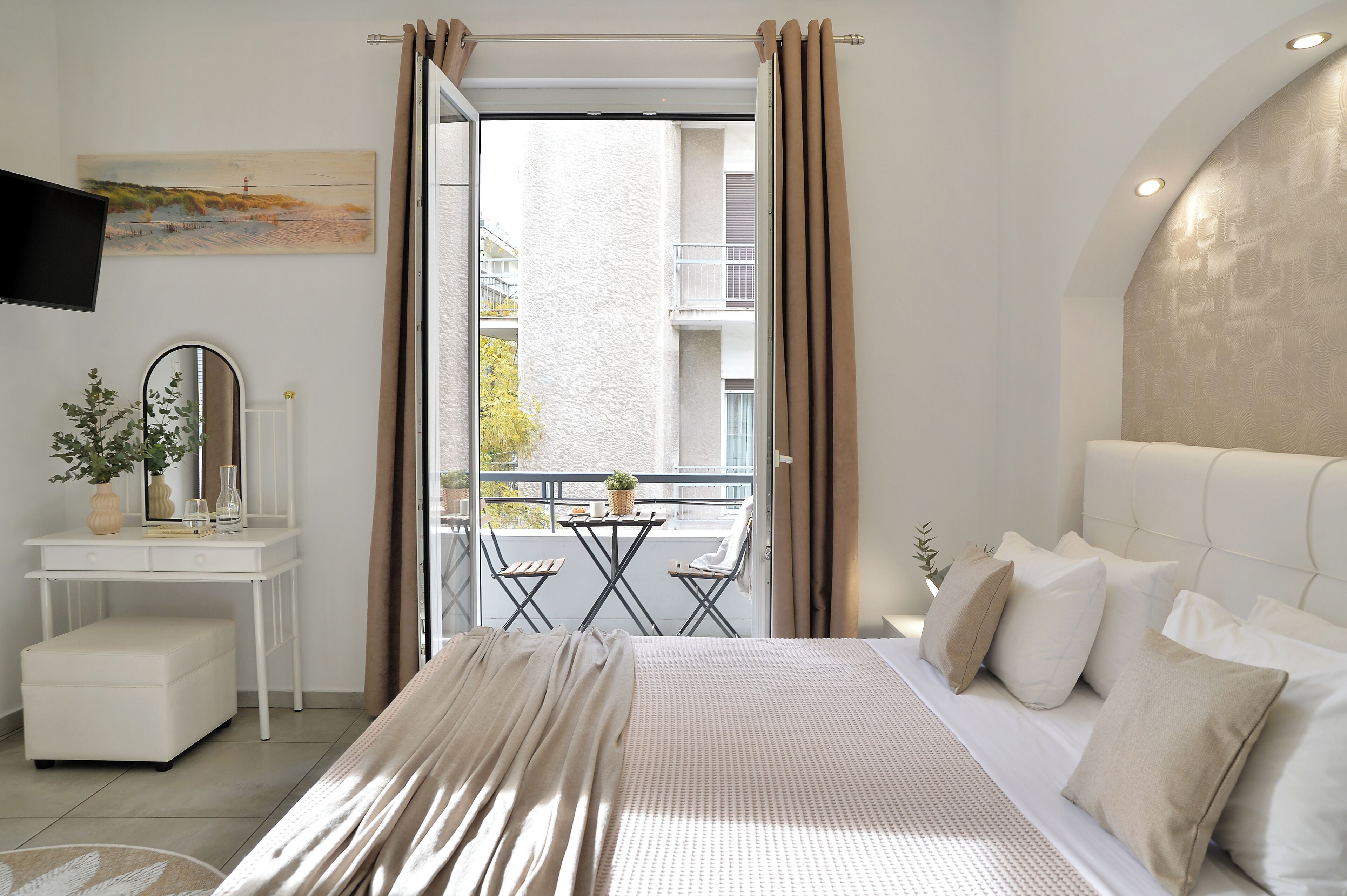 A bedroom with a bed, a bedside table with a mirror, a small table with chairs, and a balcony with a view of buildings.