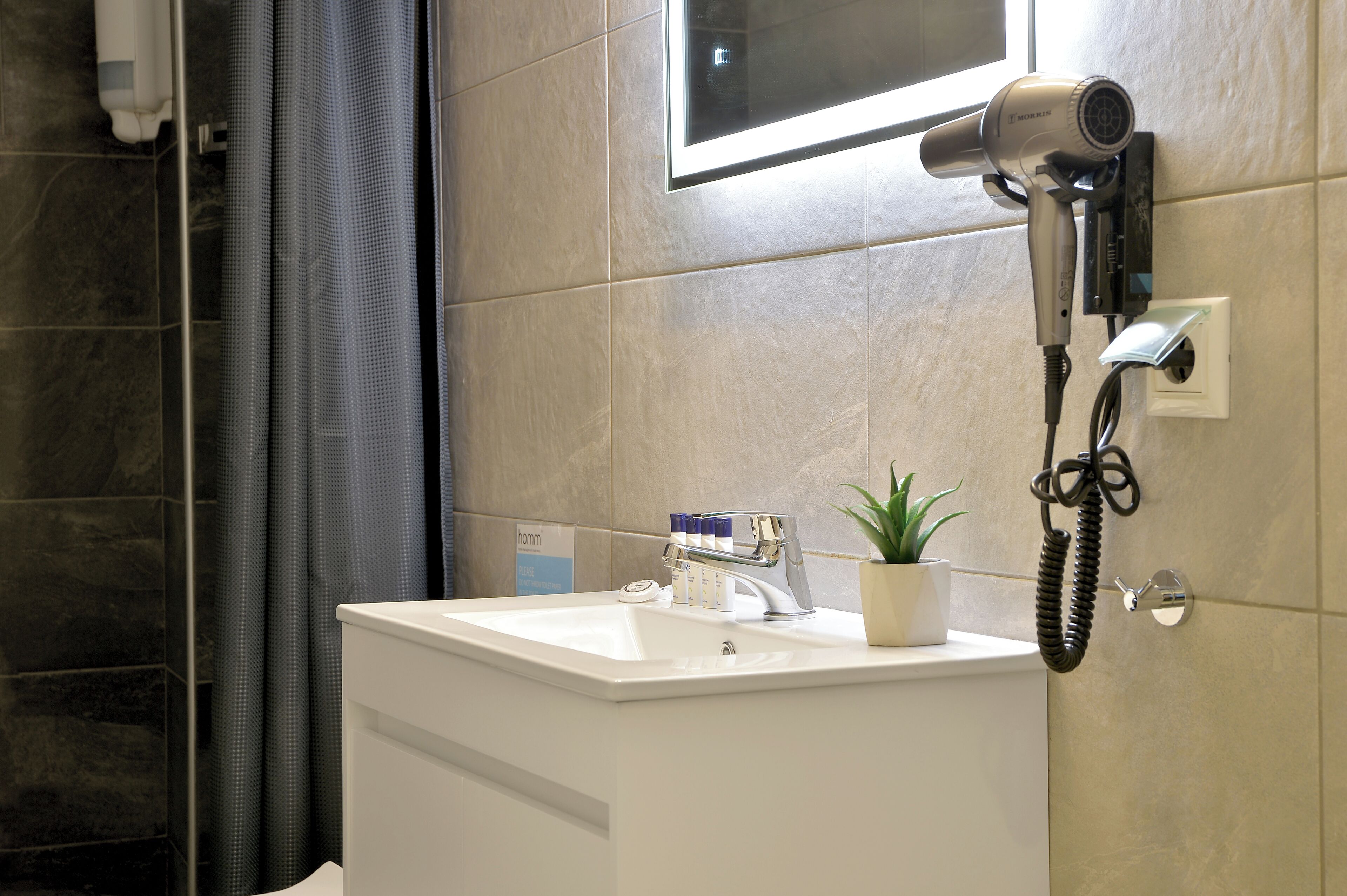 Een badkamer met een wit wastafelmeubel, een haardroger bevestigd aan de muur, een potplant en een douchehoek.