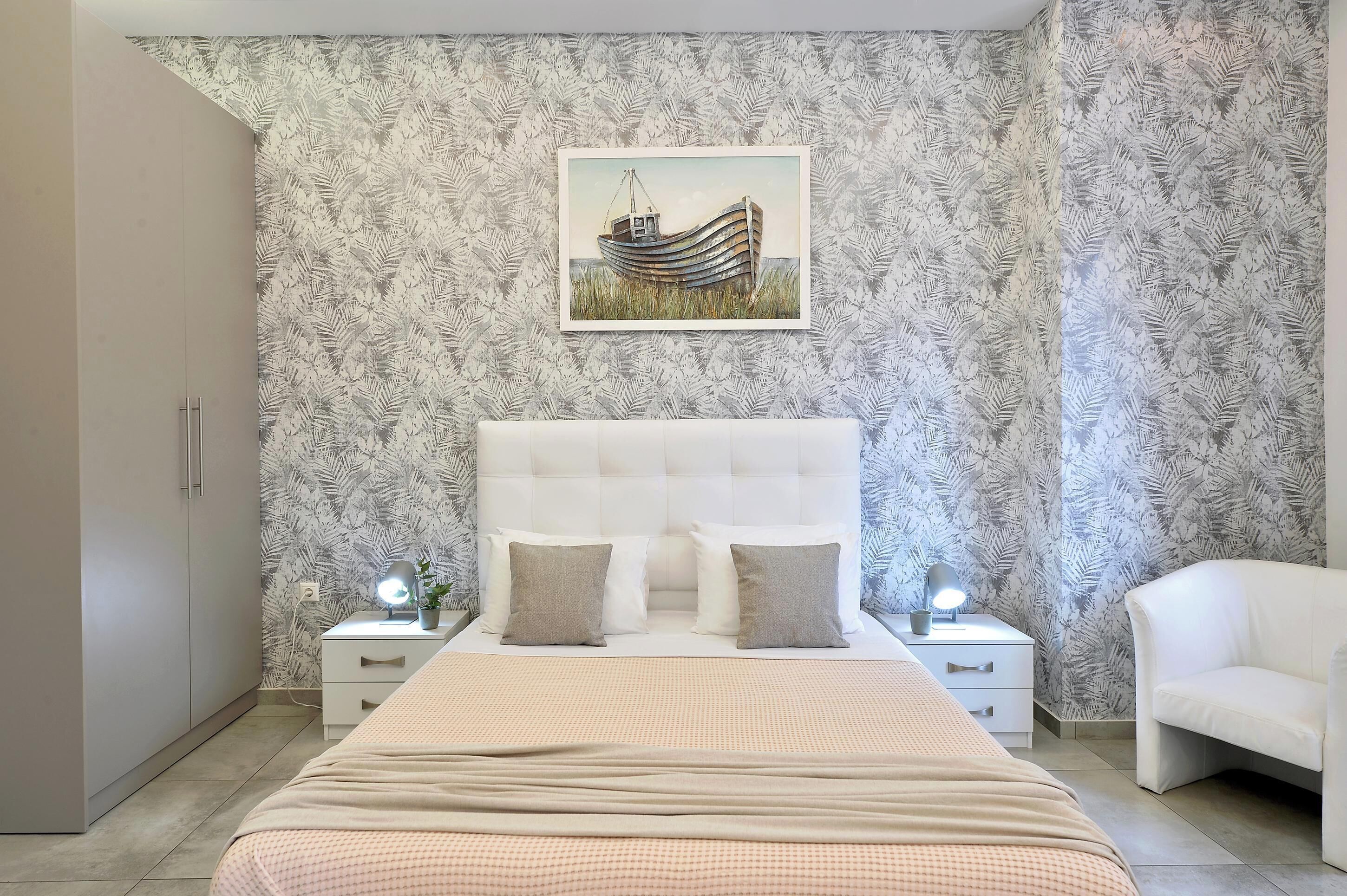 A bedroom with a bed, nightstands, a white chair, a headboard, and a wall with a leaf-patterned wallpaper.