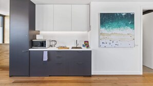 A modern kitchen with a microwave, kettle, and a coffee maker on the counter. There is a large painting of a beach scene on the wall. - Stylish Studio with Great Amenities near Beachside (St Kilda West)