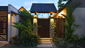 Una casa moderna con tetto scuro, parete in pietra e porta in legno. Piante e un vialetto in pietra conducono alla porta.