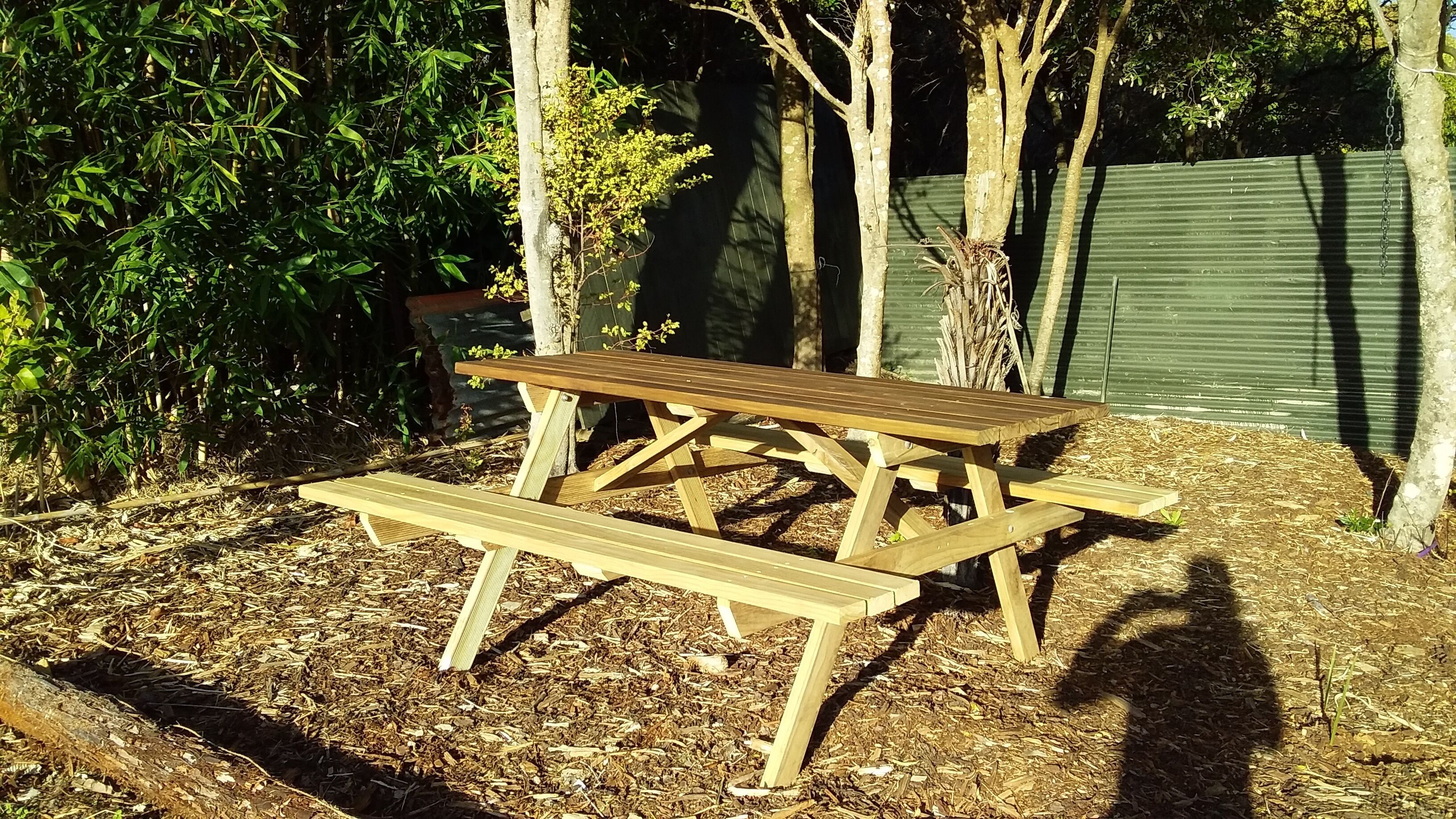 A wooden picnic table with benches in a garden area with mulch ground cover and trees in the background.
