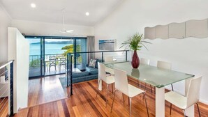 Un espace repas moderne avec une table en verre et des chaises blanches, une grande fenêtre donnant sur la mer et un escalier menant à un balcon.