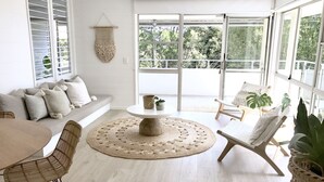 A bright, airy room with a white sofa, a round wooden table, and a chair. There are large windows and a view of greenery outside. - The Nest - 5 minute walk to Hasting Street and Noosa Main Beach (Noosa)