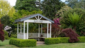 Ein Gazebo mit weißem Gestell und Dach, umgeben von gepflegten Hecken und verschiedenen blühenden Pflanzen.