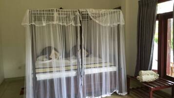 A room with a bed under a mosquito net, a bench with towels, and a window with curtains.