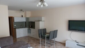 Een moderne woonkamer met een bank, een glazen eettafel met stoelen, een flatscreen televisie aan de wand en een keuken op de achtergrond.