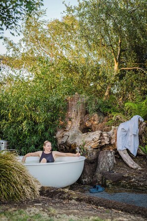 A person in a bathtub outdoors surrounded by trees and a large log.