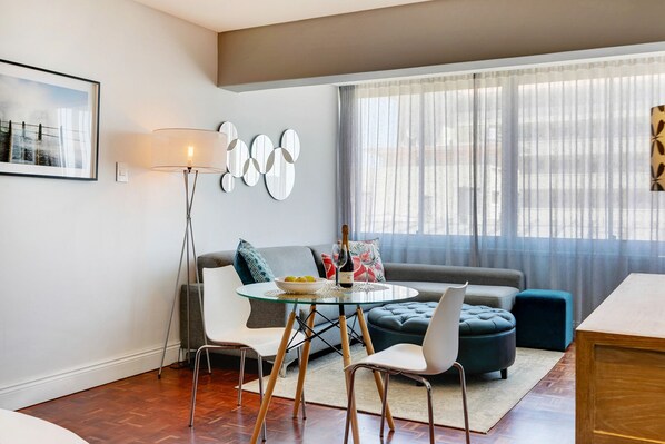 A modern living room with a glass coffee table, white chairs, a grey sofa, and a blue ottoman. There is a floor lamp, a framed picture on the wall, and a window with sheer curtains.