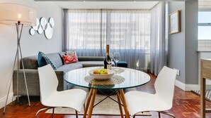 A modern living room with a glass dining table, white chairs, a grey sofa, and a floor lamp. - Backup-Power Perfect Modern Beach Apartment near V&A with Sea and Park Views (Cape Town)