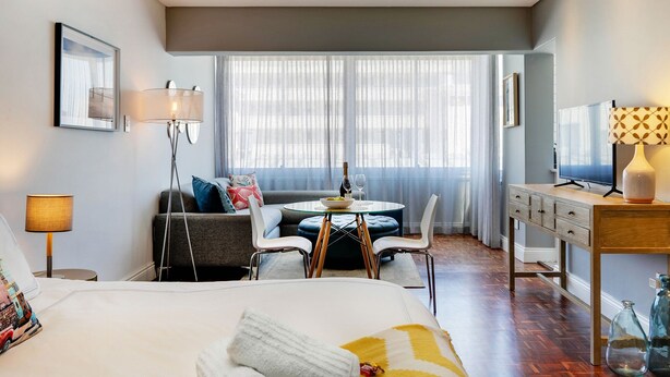 A modern hotel room with a bed, a dining table, a sofa, and a TV. - Backup-Power Perfect Modern Beach Apartment near V&A with Sea and Park Views (Cape Town)