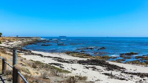 A coastal beach with rocky shoreline, clear blue water, and a distant ship. - Backup-Power Perfect Modern Beach Apartment near V&A with Sea and Park Views (Cape Town)