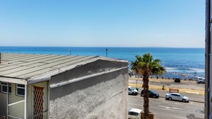 A view of a beach with a clear sky, a palm tree, and parked cars along the shore. - Backup-Power Perfect Modern Beach Apartment near V&A with Sea and Park Views (Cape Town)