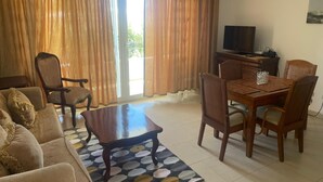 A living room with a sofa, coffee table, dining table with chairs, a TV, and a view through the curtains.