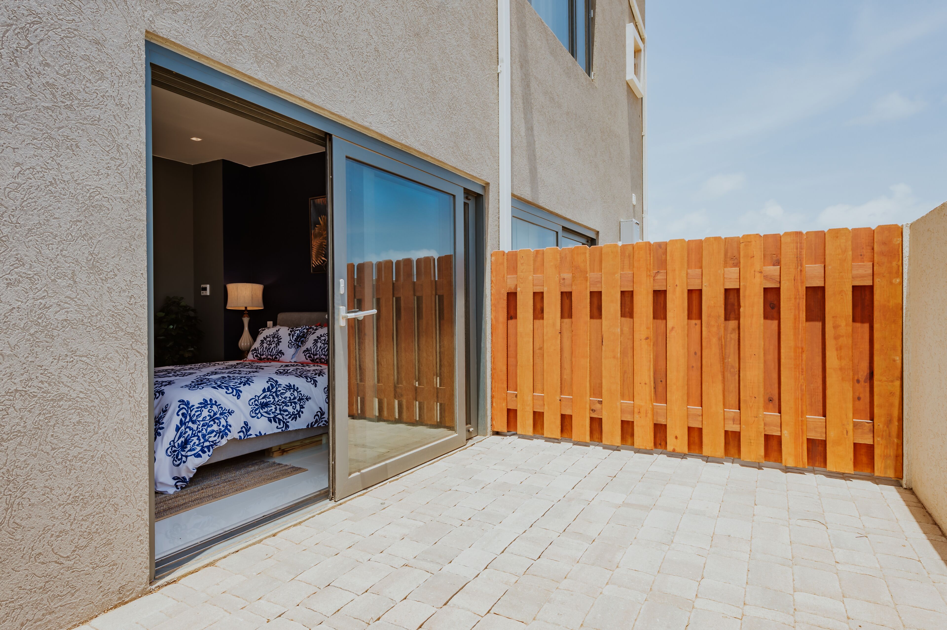 A bedroom with a sliding glass door, a bed with a patterned bedspread, and a wooden fence in the background.