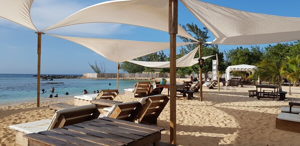 Une plage avec des chaises longues et des tables en bois, abritées sous des auvents blancs, une clôture en bois et une vue dégagée sur la mer.