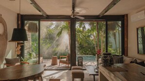 A spacious room with a large sliding glass door leading to a tropical garden, a ceiling fan, and a wooden dining table. - Secluded Two-Bedroom Beach House (Tulum)