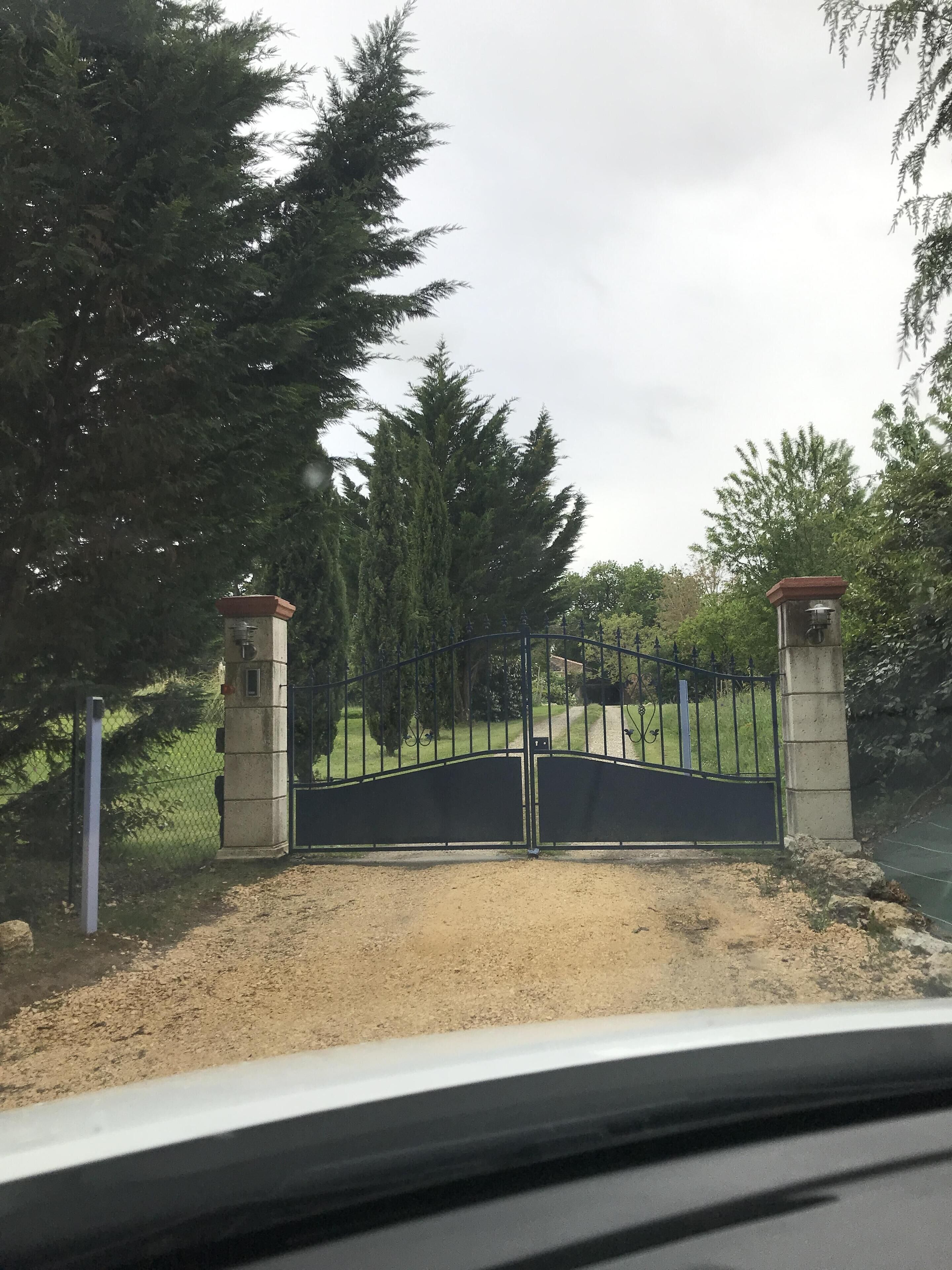 A gated entrance with a metal gate, flanked by two stone pillars, leading to a grassy area with trees.