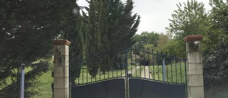 A gated entrance with a metal gate, flanked by two stone pillars, leading to a grassy area with trees.