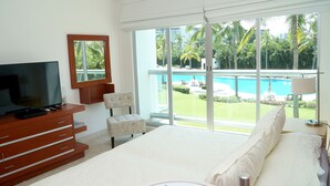 A bedroom with a large bed, a TV on a wooden dresser, a chair, and a view of a swimming pool and palm trees. - Súper Condo 3 at Vidanta (Nuevo Vallarta)