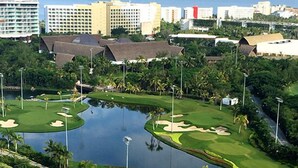 A golf course with a water hazard, surrounded by palm trees and a view of a city skyline in the background. - Súper Condo 3 at Vidanta (Nuevo Vallarta)