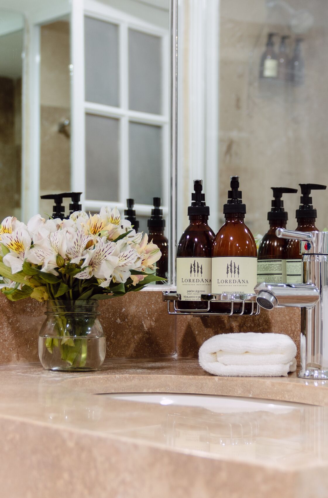 A bathroom vanity with a vase of flowers, a white towel, and several Loredana skincare bottles.